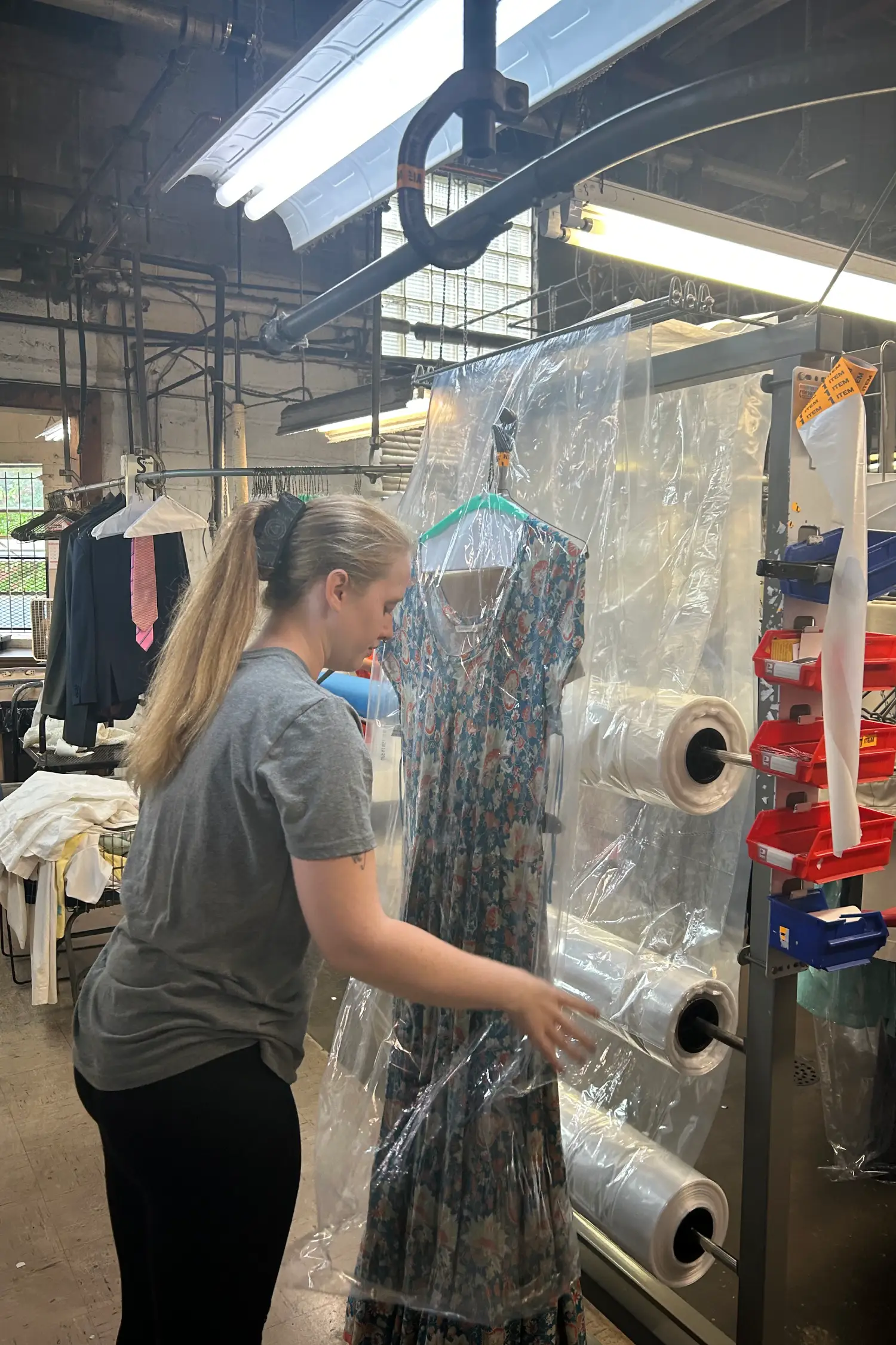 A woman in a gray shirt inspects a floral dress hanging behind a plastic barrier in a laundry facility, surrounded by clothing items.