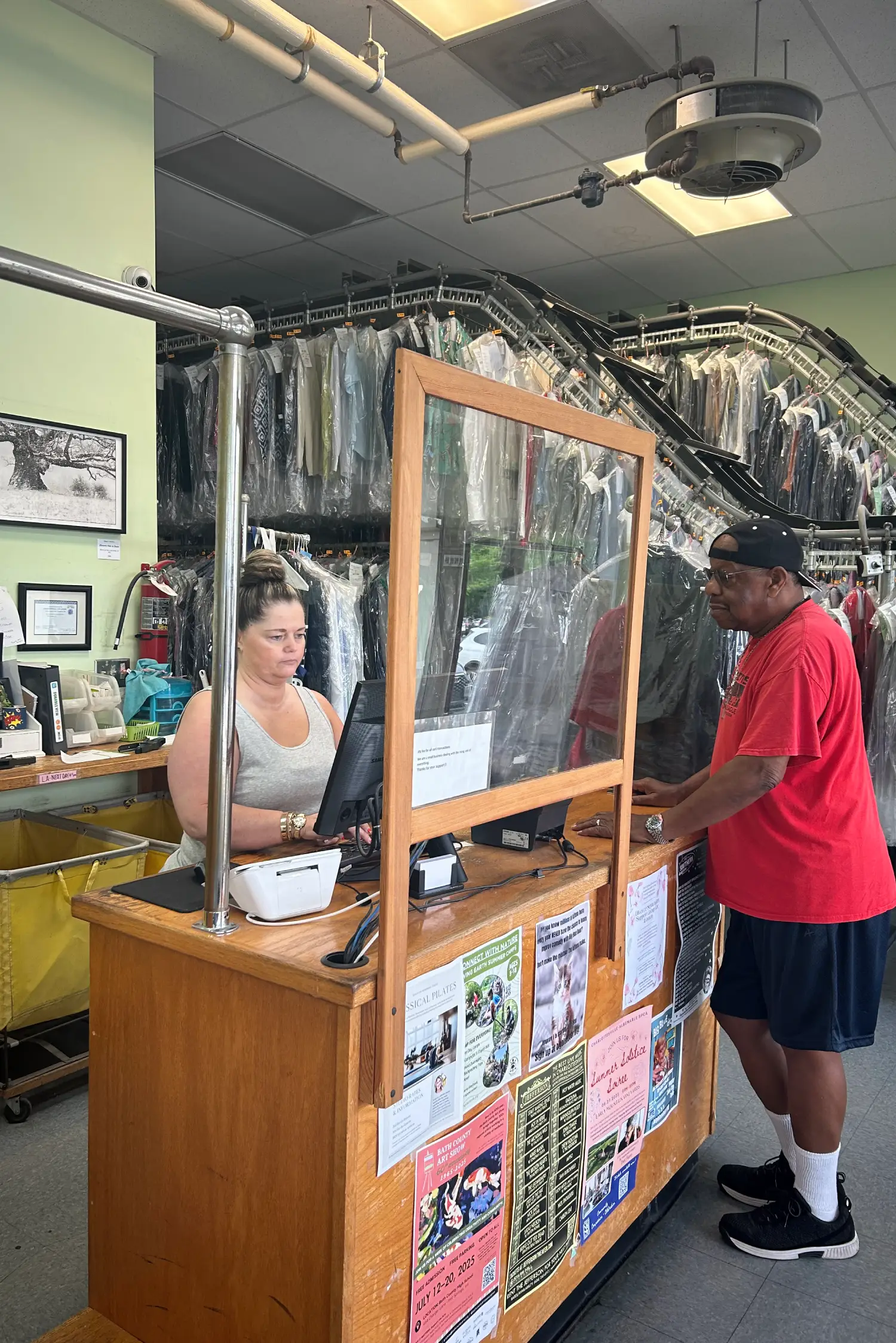A customer in a red shirt interacts with a worker at a wooden counter surrounded by hanging garments in a clean laundromat.