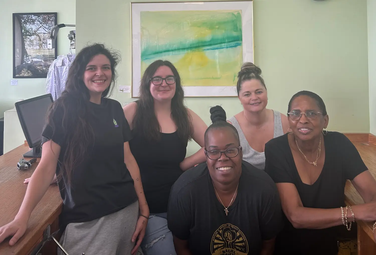 A group of five women poses in front of a green and yellow abstract painting, with a wooden countertop and computer equipment nearby.